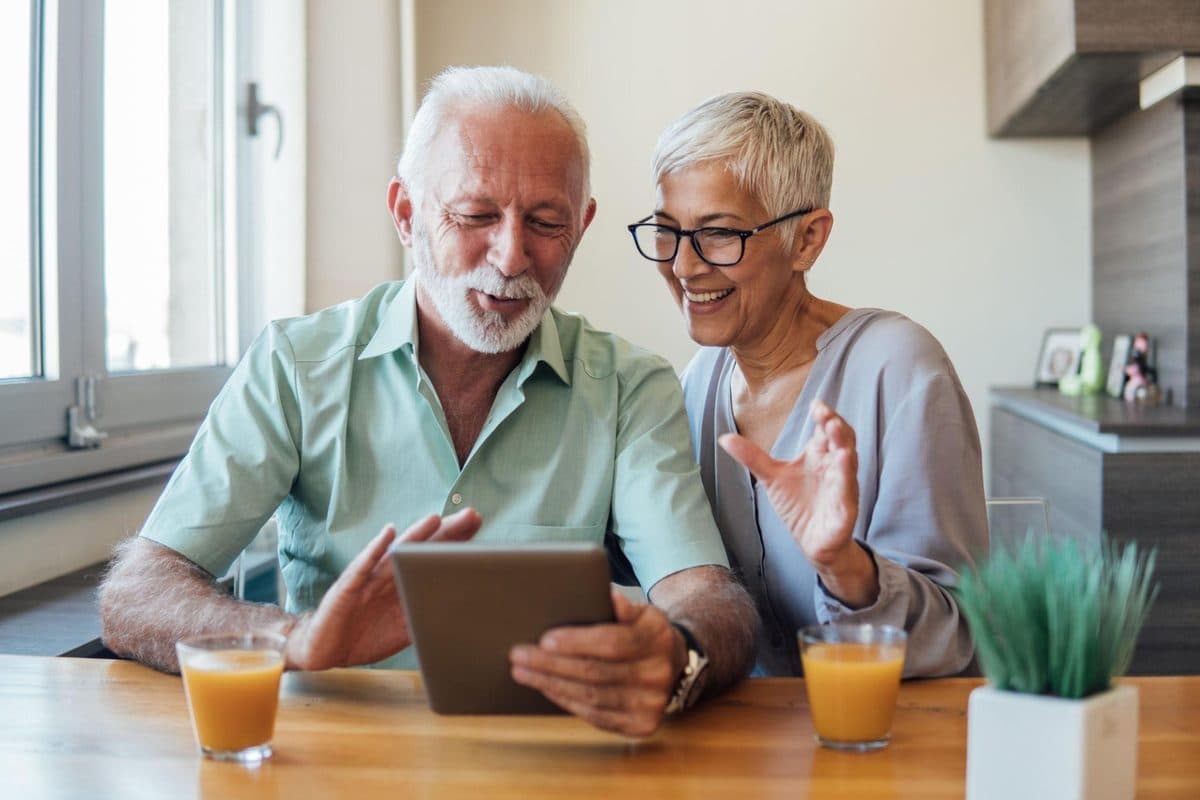 Een oudere man en vrouw kijken samen glimlachend op een tablet aan de keukentafel